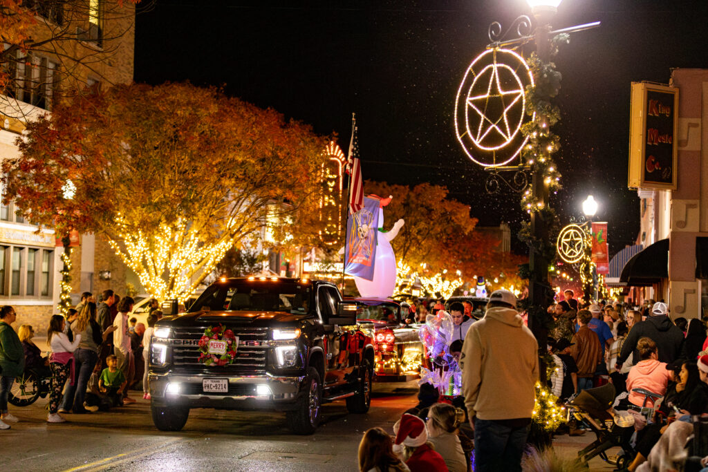 Image of center avenue in downtown Brownwood with parade truck driving past with crowds on the sidewalks and the trees and vehicles are lighted with christmas lights.